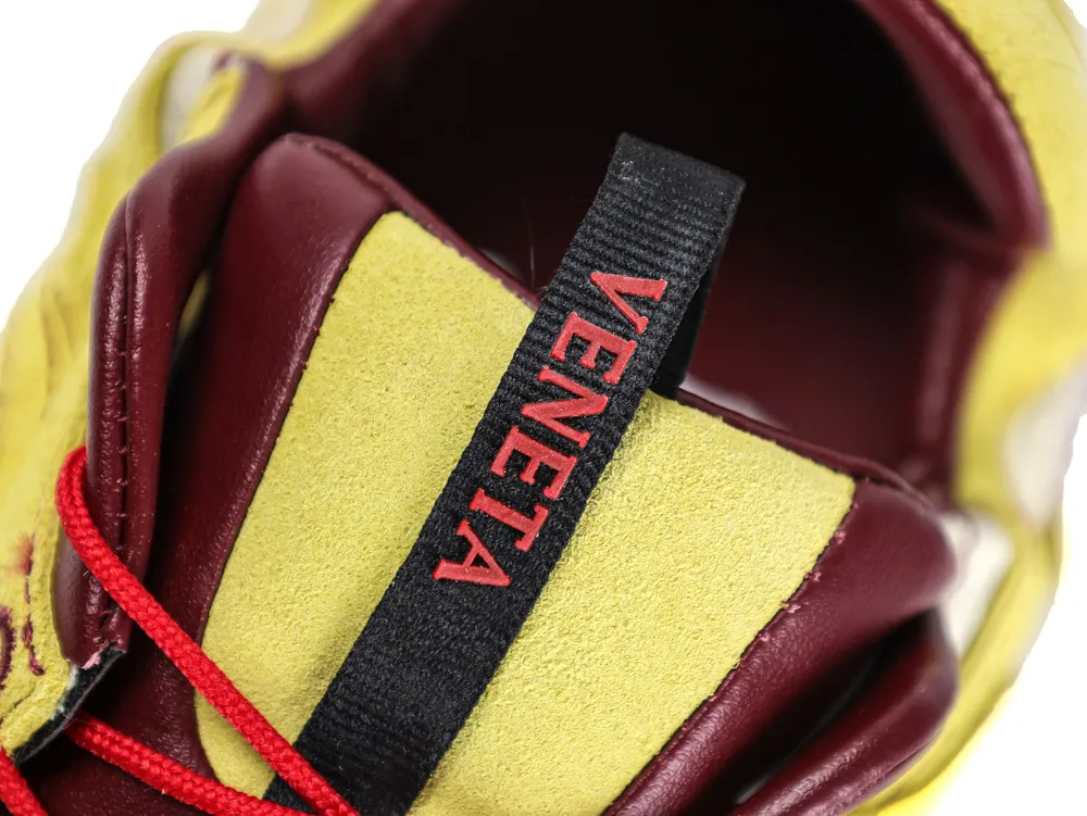 Close-up of a sneaker with yellow suede, red leather, and black fabric pull tab.