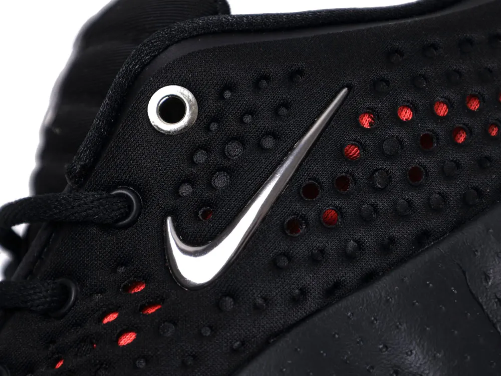 Close-up of black fabric shoe with red perforations and a silver logo.