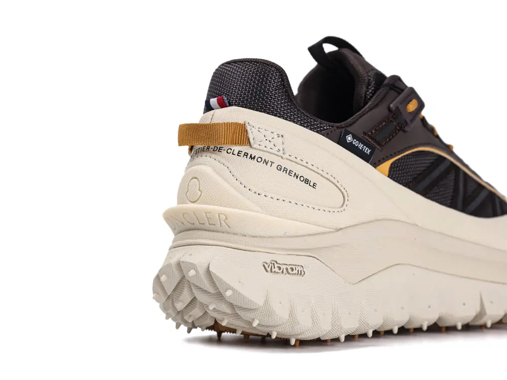 Close-up of a black and beige sneaker featuring Gore-Tex technology, detailed stitching, and textured sole. Labels include Moncler and Vibram. The shoe has a pull tab and design elements for outdoor use.